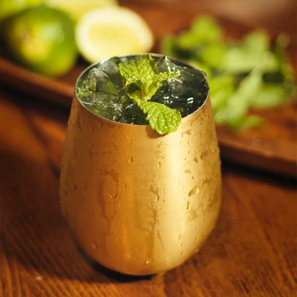 A golden cup filled with ice and a mint sprig, with limes and mint in the background.