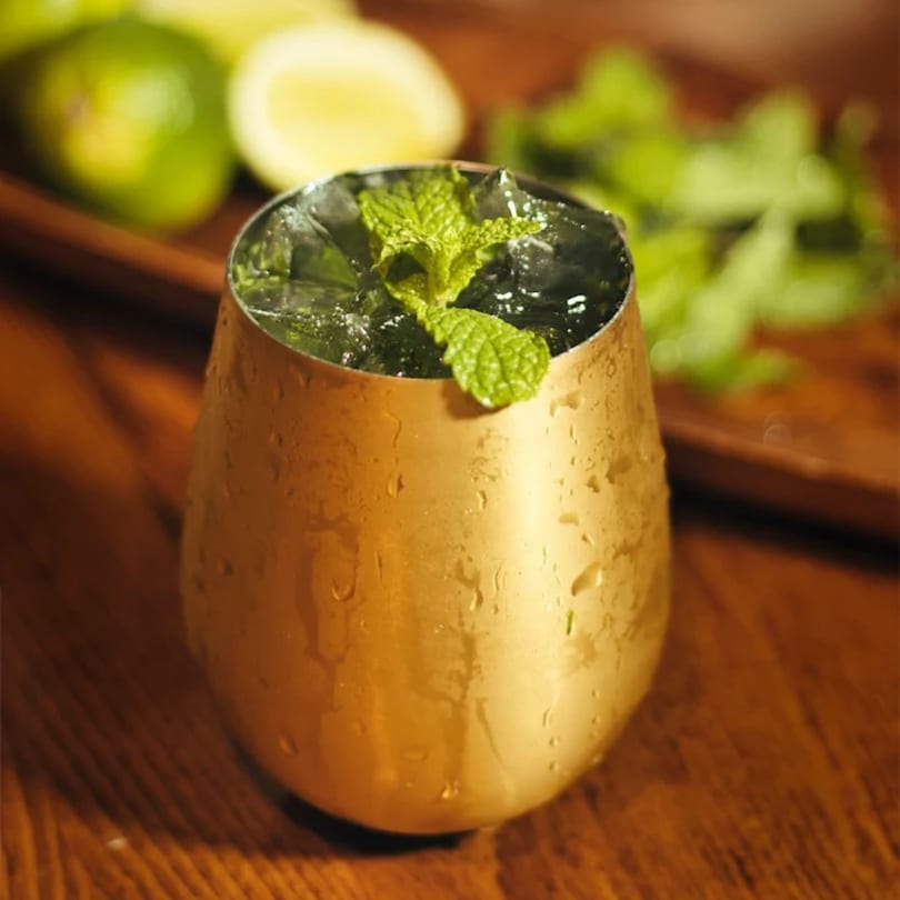 A golden cup filled with ice and a mint sprig, with limes and mint leaves in the background.